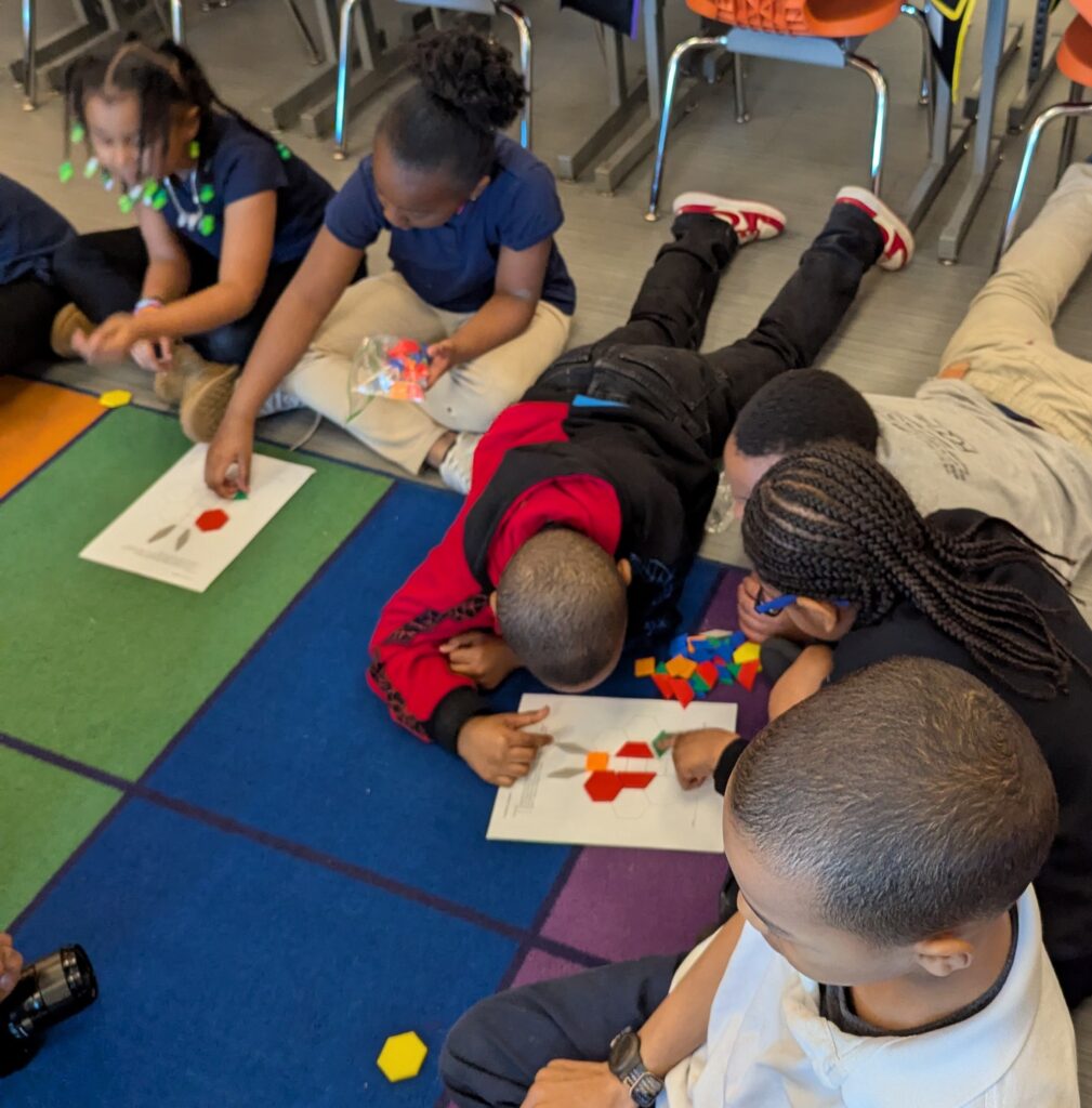 A group of young students sitting and lying on a colorful classroom carpet, working together with pattern blocks and activity sheets.