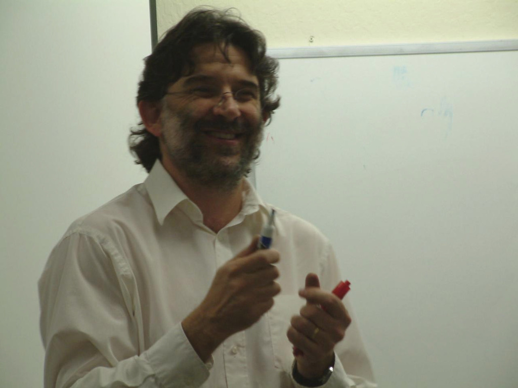 Bill McCallum smiles while holding markers in a classroom. He wears a white dress shirt and glasses and stands in front of a whiteboard.