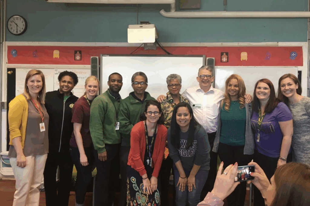 Bill McCallum stands with a group of educators and staff in a classroom. They pose together in front of a whiteboard and red bulletin board, smiling for a group photo.