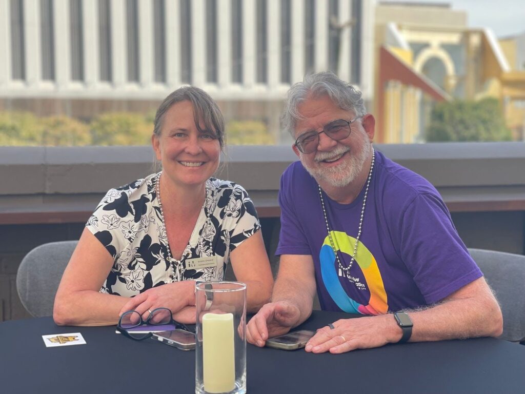 Bill McCallum and Kristin Umland sit at an outdoor table, smiling. He wears a purple Illustrative Mathematics shirt and beads; she wears a black-and-white floral top. A tall candle is on the table.